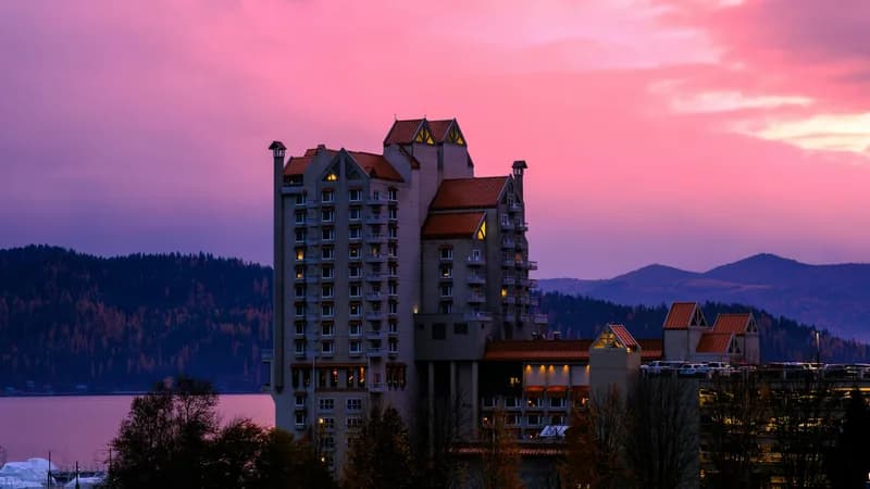 The Coeur d'Alene Resort tower lit from within at dusk, silhouetted against a pink and violet sunset over the lake and distant mountains.