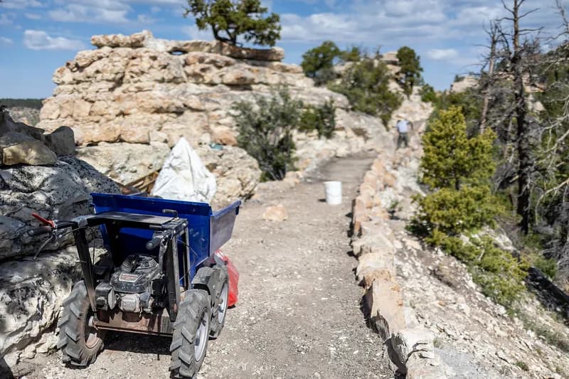 A powered wheelbarrow and bagged trail material parked on a freshly rebuilt canyon-rim path, with limestone outcrops and junipers on either side.