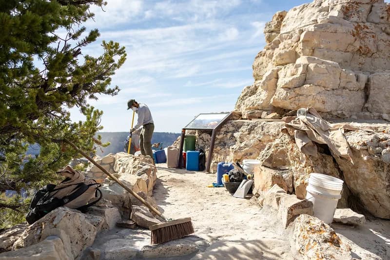 A trail worker raking fresh tread at a canyon-rim work site, surrounded by buckets, hand tools and stacked stone.