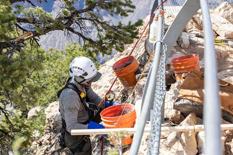 A worker in a climbing helmet and harness hauling orange buckets along a rigged line on a steep, rocky canyon slope.