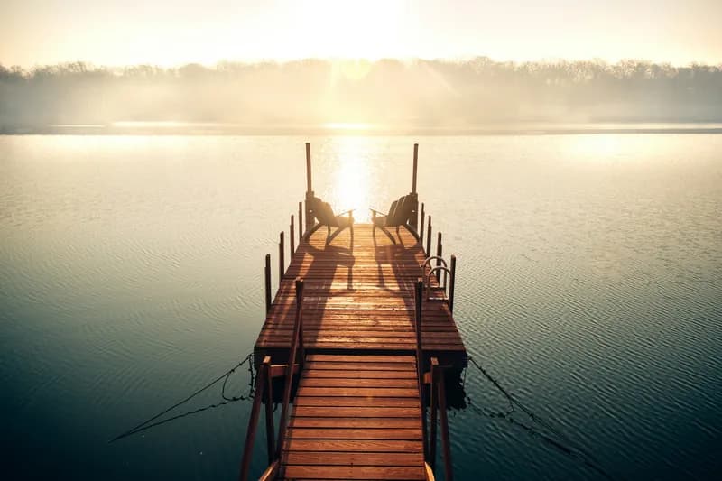 A wooden dock running out to two empty chairs at its end, backlit by golden mist rising off a calm lake at sunrise.