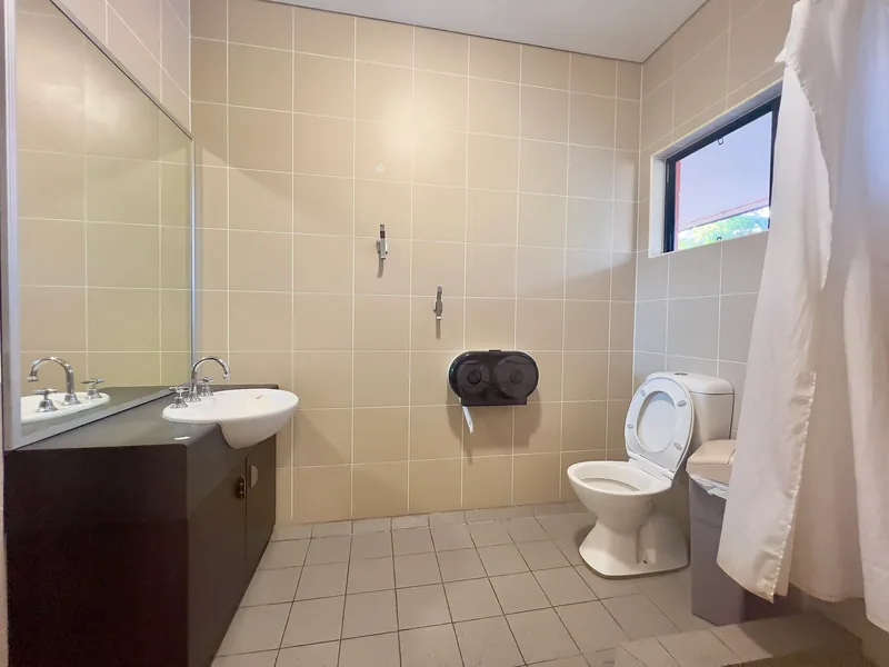 A simple hostel bathroom with a sink, wall mirror, toilet, tiled shower floor, and white shower curtain.