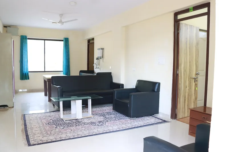 A bright hostel common area with black sofas and armchairs arranged around a glass coffee table near a large window.
