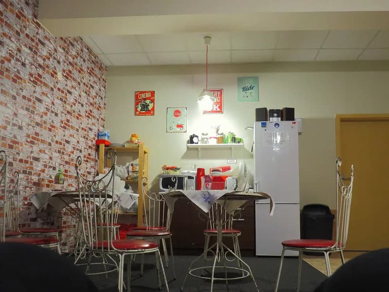 A shared hostel kitchen with a refrigerator, dining table, sink, microwave, and open shelving against a brick-patterned wall.