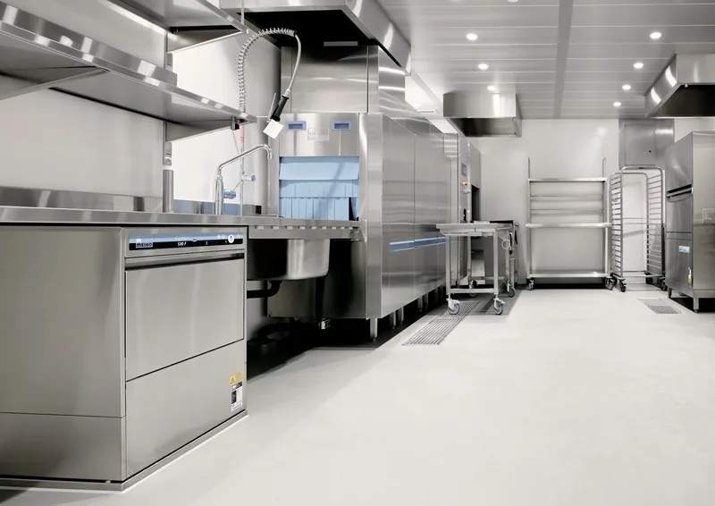 An empty commercial kitchen in stainless steel, with a pass-through dishwasher, deep sinks, prep tables and sheet-pan racks under bright ceiling lights.