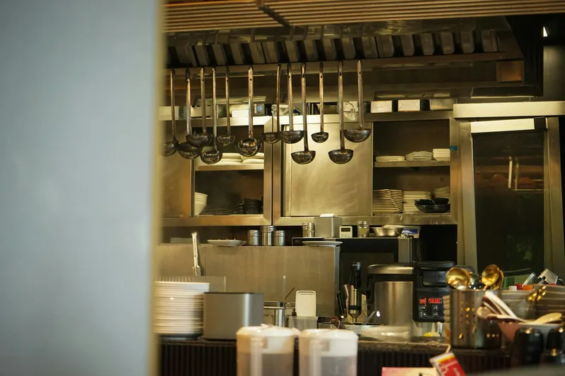 A row of ladles hanging above a restaurant service pass, with stacked white plates, bowls and prep containers on the line behind.