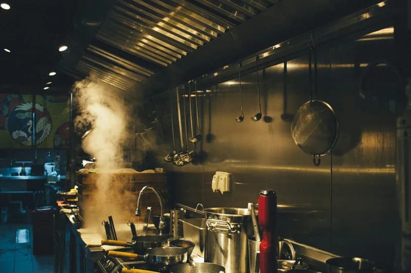 Steam rising off a wok on a dark restaurant cook line, with utensils and a strainer hanging along a stainless steel wall.