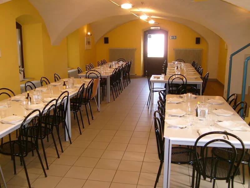 An empty youth-hostel dining room set with long white tables, place settings, and rows of black chairs beneath a vaulted ceiling.