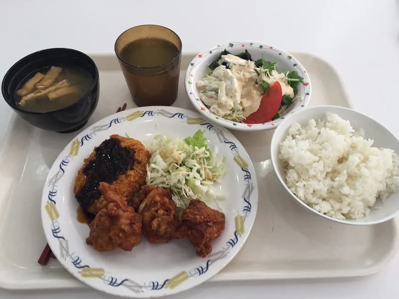 A cafeteria lunch tray with rice, fried chicken, a breaded cutlet, shredded cabbage, salad, soup, and tea.