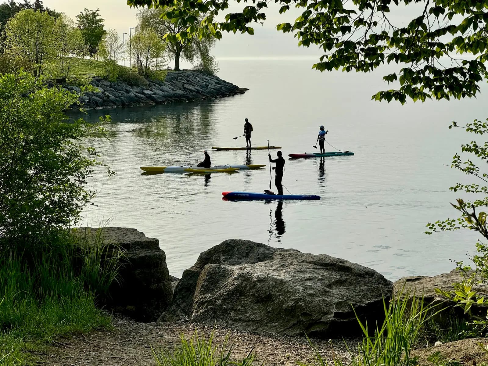 Paddleboarders and kayakers silhouetted on calm water at dusk, seen from a rocky, tree-framed shoreline.