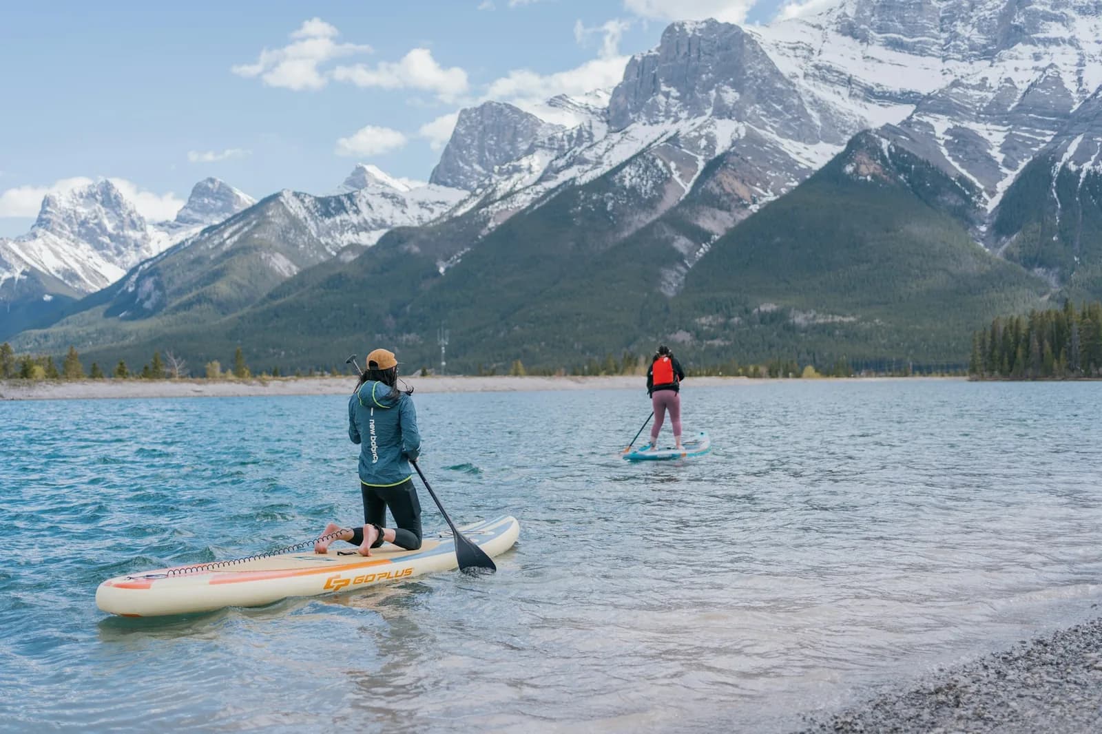 Two paddleboarders seen from behind, gliding across a shallow turquoise lake toward snow-covered mountain peaks.