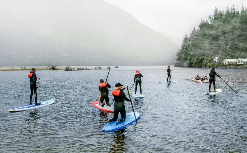 A group of paddleboarders in wetsuits and life vests spread across a misty lake below forested hills.